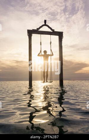 Young man standing on rope swing at beach against sunset, Gili Trawangan, Lombok, Indonesia Stock Photo