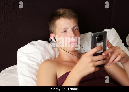 A teenager lies in bed with headphones and a smartphone in his h Stock Photo