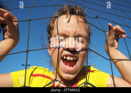 An 8-year-old Caucasian girl stands behind a sports net and screams. Volleyball sports dividing net against the blue sky close-up. Outdoor sports Stock Photo