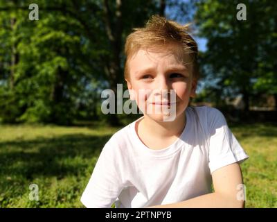 Blond boy 7 years old in a white T-shirt on the background of a forest. The child looks to the side, squints his eyes from the bright sun. Portrait on Stock Photo