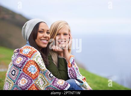 Friendship keeps us warm. Two young female friends sitting wrapped up in a blanket outside Stock Photo