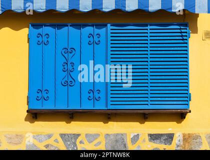 Architecture of an old yellow wall with an old blue metal window outside. Exterior texture details of an old rustic residential build Stock Photo