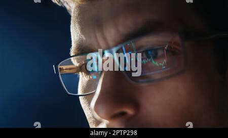 Close up view of focused trader wears eyeglasses looking at pc screen with stock market charts reflection, working online late night. Man analysing crypto market. Selective focus on eye. Copy space.  Stock Photo