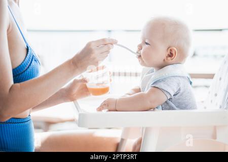 Mother spoon feeding her baby boy child in baby chair with fruit puree on a porch on summer vacations. Baby solid food introduction concept Stock Photo