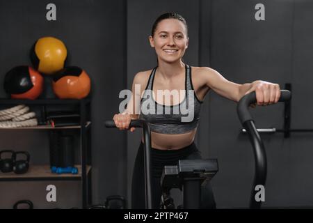 Portrait of smiling fitness woman using air bike for cardio workout and exercising her legs at gym. Happy crossfit female athlete training on sport equipment in fitness club. Cycling training Stock Photo