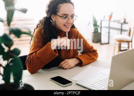Happy student learning online from home, taking notes while watching webinar on laptop. Smiling young woman attending virtual meeting on laptop while Stock Photo