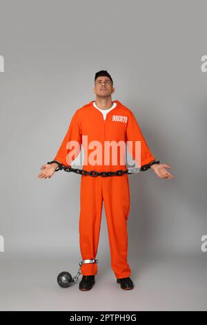 Prisoner in orange jumpsuit with chained hands and metal ball on grey background Stock Photo