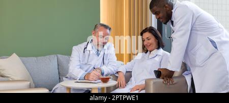 Group of doctors in white gown and stethoscope sit relax on sofa during coffee break. Senior professor with mustache and beard writing on clipboard sh Stock Photo