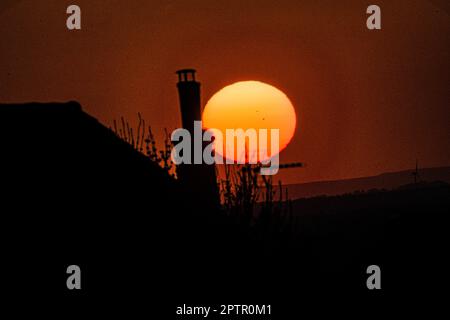 A beautiful bright orange English spring sun set with a silhouette of a chimney and television aerial in the foreground Stock Photo