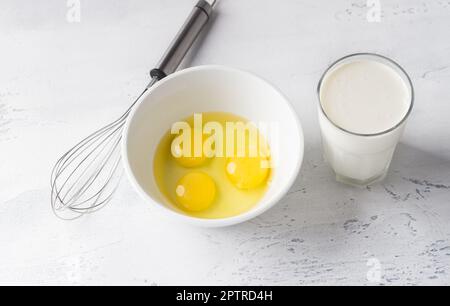 White bowl with three broken eggs without shell, a glass of cream or milk and a whisk for whisking on a light gray background, top view. Cooking homem Stock Photo