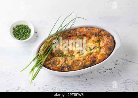 Casserole or clafoutis with mushrooms and cheese in a white ceramic baking dish sprinkled with green onions on a light background. Delicious homemade Stock Photo