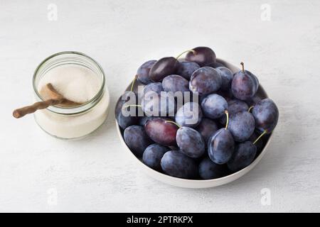 A bowl of plums and a jar of sugar, ingredients for making plum jam on a light gray background Stock Photo