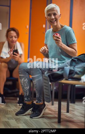 Mature senior woman in sportswear sitting on bench in gym locker room, smiling holding an apple and feeling ready for workout. Stock Photo