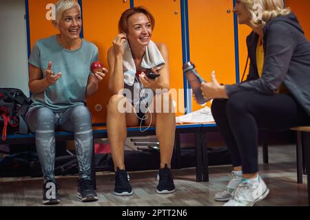 Two mature senior women and younger woman in sportswear sitting in locker room after training, having conversation, enjoying time together. Stock Photo