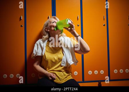 Beautiful senior blonde woman sitting in gym locker room drinking water after workout. Health, senior life, lifestyle concept. Stock Photo
