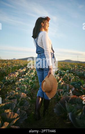 Woman farmer standing in a cabbage field on a farm. Young female with a straw hat and rubber boots looking over her field of organic vegetables. Stock Photo