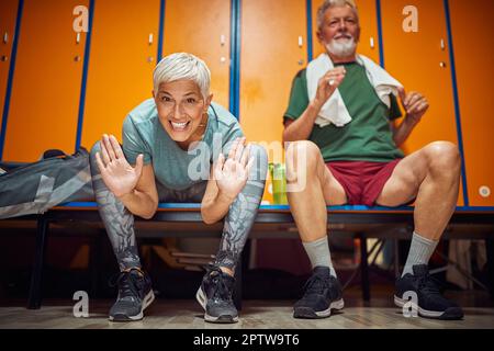 Joyful senior couple sitting in gym locker room, feeling energized and acting goofy, pre workout. Health, togetherness, active lifestyle concept. Stock Photo