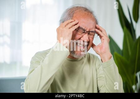 Headache. Close up of elderly holds head with hand suffering from migraine headache, Sad Asian senior man sitting on sofa feeling hurt and lonely at h Stock Photo