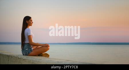 Calm young woman in tank top and denim shorts sits in lotus position on the pier at sunset Stock Photo