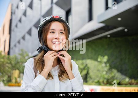 Asian young businesswoman putting biking helmet prepared cyclists around building go to work, Close up smiling woman wearing helmet exercise outdoors Stock Photo