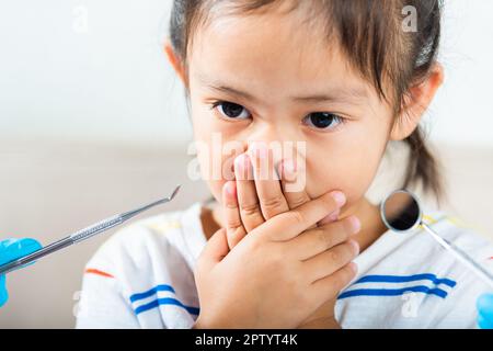 Dental kid examination. Doctor examines oral cavity of child uses mouth mirror to check teeth cavity but little girl cover mouth no need to checking s Stock Photo