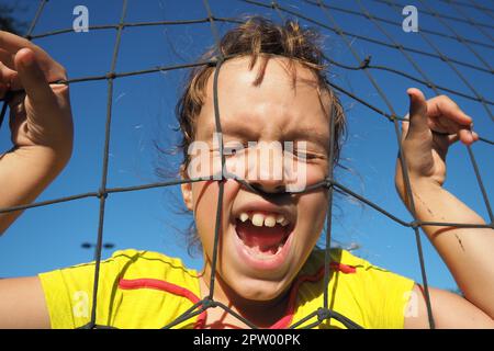 An 8-year-old Caucasian girl stands behind a sports net and screams. Volleyball sports dividing net against the blue sky close-up. Outdoor sports Stock Photo