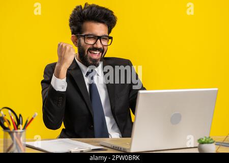 Indian businessman working on laptop at office desk typing browsing say Wow yes found out great big lottery jackpot win, good job news, celebrating. Freelancer arab man isolated on yellow background Stock Photo