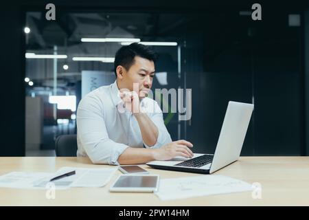 Serious and thoughtful young man of Asian origin programmer, developer works in office at desktop at laptop. Stock Photo