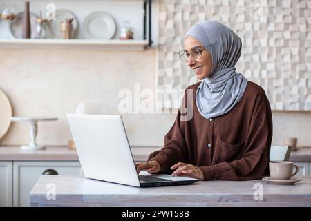Muslim young woman in hijab programmer, developer working at home, sitting in kitchen at laptop. Stock Photo