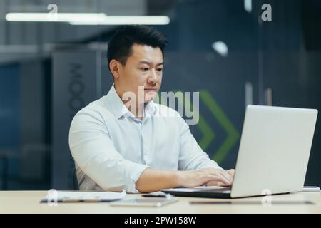 A young man of Asian origin programmer, developer works in the office at a desktop using a laptop. Stock Photo