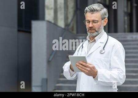 Portrait of a senior gray-haired male doctor standing in a white coat outside the clinic and using a tablet. Stock Photo