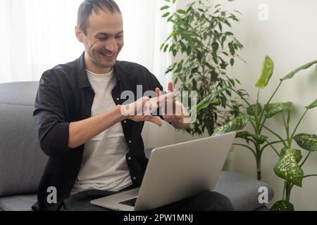 Happy smiling deaf young caucasian man uses sign language while video call using laptop while sitting at home, virtual communication concept. Stock Photo