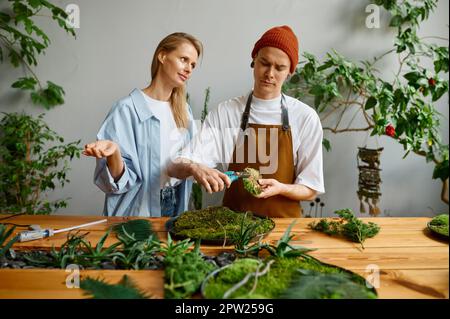 Florist and decorator team working at DIY florarium studio. Young woman criticizing hipster man while he cutting moss piece with scissors Stock Photo