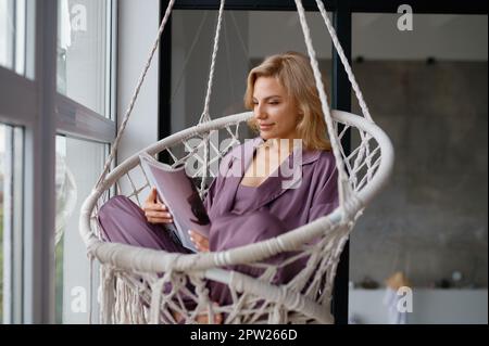Woman rest in lounge hanging wicker chair at home. Glazed balcony cozy house interior Stock Photo