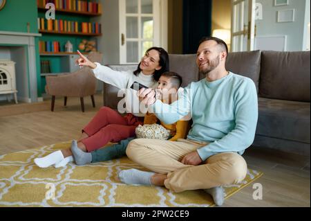 Amazed parents and curious kid watching tv program sitting on floor in living room at home Stock Photo