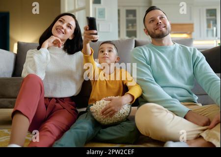 Amazed parents and curious kid watching tv program sitting on floor in living room at home. Close up view Stock Photo