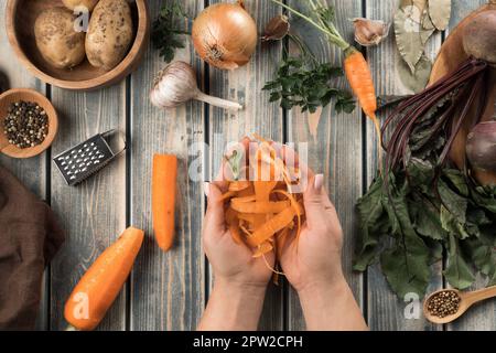 Unrecognizable woman holding carrot peelings in hands near wooden bowl with potatoes, little bowl with peppercorns, carrots, onion, garlic, beets on t Stock Photo