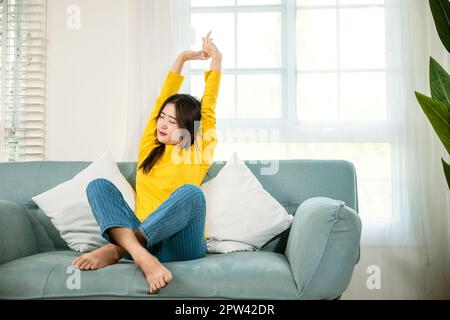 Lifestyle Asian young woman stretching her arms on sofa in the living room at home, happy female relaxing activity feeling lazy on weekend on couch, r Stock Photo