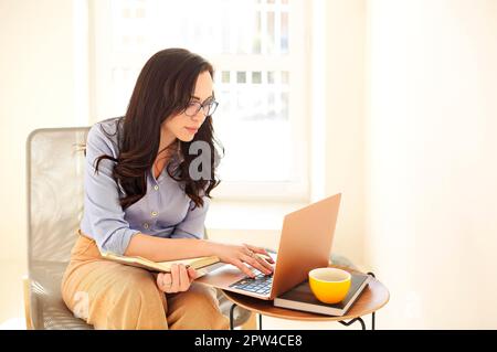 Confident elegant woman in glasses sitting in armchair with laptop on small table aside and taking notes in planner Stock Photo