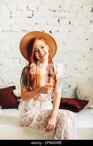 Happy young female in boho dress and hat enjoying wine and looking at camera with smile while sitting on bench against white brick wall Stock Photo