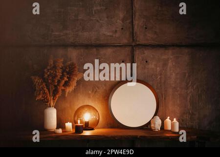 Round mirror on wooden shelf with two burning candles and small porcelain Buddha head reflected in corner of mirrored surface over concrete wall Stock Photo