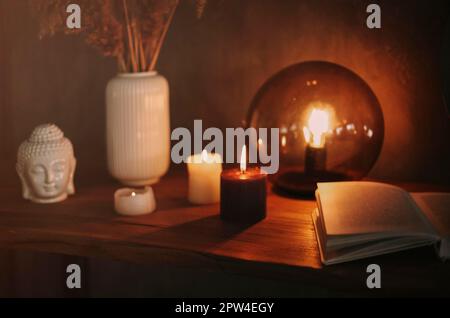 Round mirror on wooden shelf with two burning candles and small porcelain Buddha head reflected in corner of mirrored surface over concrete wall Stock Photo