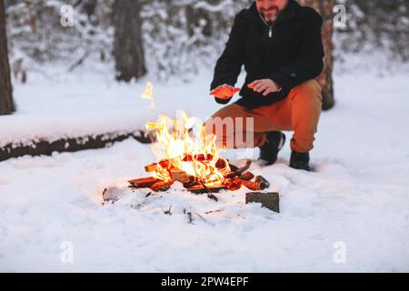 Mature man warming his hands with fire after making campfire in middle of snowy forest in cold winter evening, using heat to feel good and Stock Photo