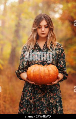 Portrait of beautiful young blond woman with long curly hair and dark vamp makeup looking down, wearing black guipure dress, holding pumpkins while Stock Photo
