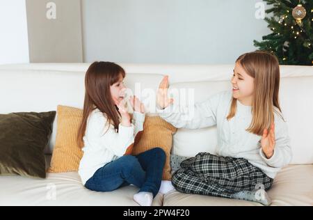 Portrait of cheerful smiling little sisters sitting on cozy couch in living room, playing clapping patty-cake game on hands, having fun, enjoying Stock Photo