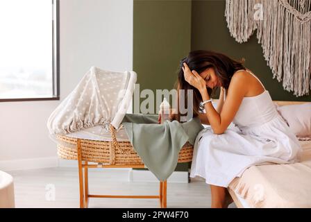 Motherhood and depression concept. Young tired mother holding milk bottle sitting near bassinet with little baby, exhausted woman mom touching head Stock Photo