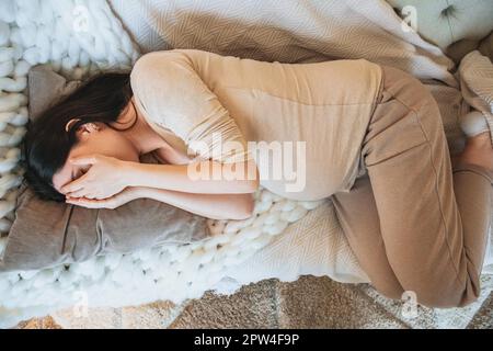 View from above of sleeping pregnant woman covering eyes with hands and relaxing on couch at home, expectant mother lying resting on sofa indoors. Stock Photo