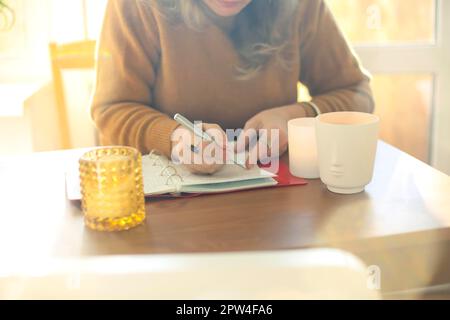 Middle-aged woman writer or poet writing in notebook while sitting at table in sunny room at home, creating novel or poetry. Focused mature female Stock Photo