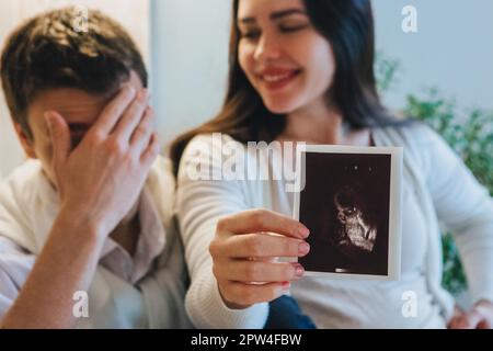 Young happy pregnant woman holding pregnancy ultrasound, showing sonogram picture to pleasantly surprised husband crying from happiness. Family Stock Photo