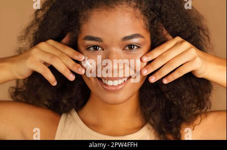 Smiling dark skinned woman with curly afro hair applies face cream on cheeks with both hands, poses over beige studio wall background, uses cosmetic Stock Photo